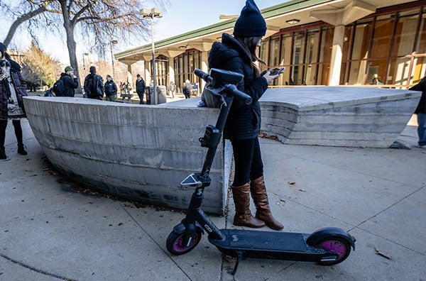 A female student is standing next to an electric scooter and using her phone outside the library